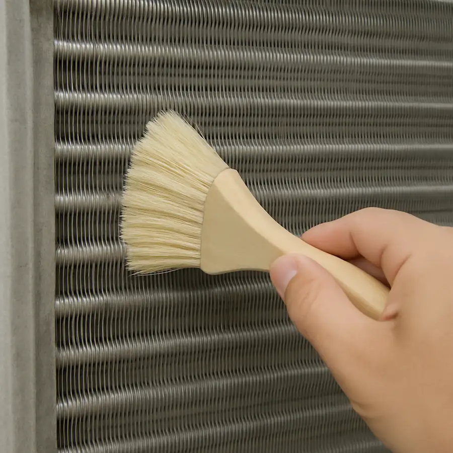 A close-up of a person using a soft brush to gently clean the fins of a metal condenser coil.