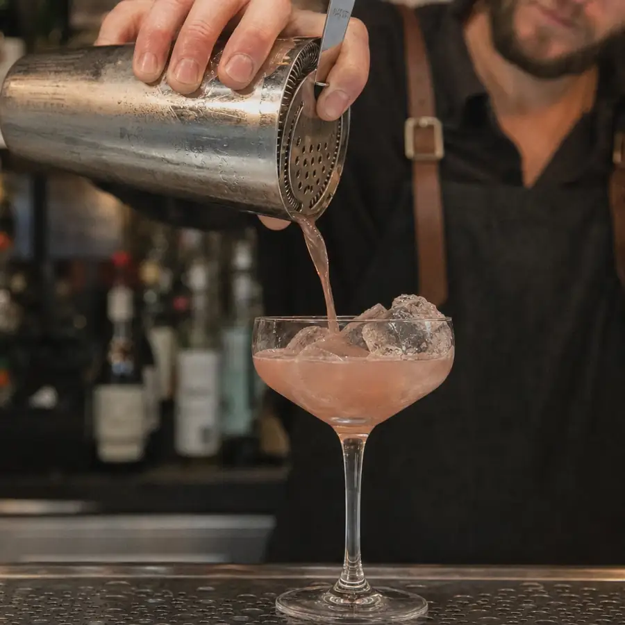 Close-up of a mixologist pouring a cocktail from a Boston shaker into a coupe glass over clear craft ice