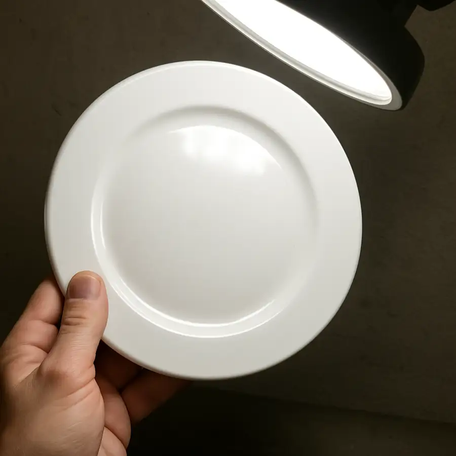 A photo showing a single white plate being inspected under a bright light for surface imperfections.