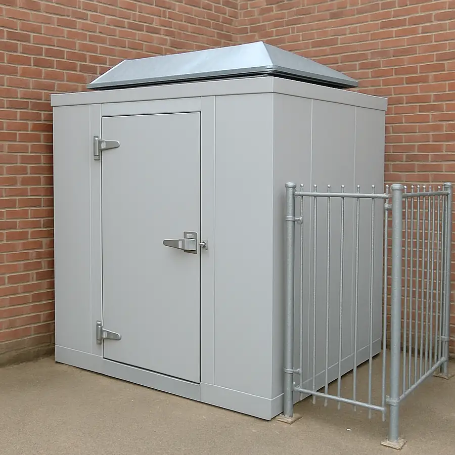 An outdoor walk-in unit installed against a brick wall with a visible weather-proof roof cap and a security fence.