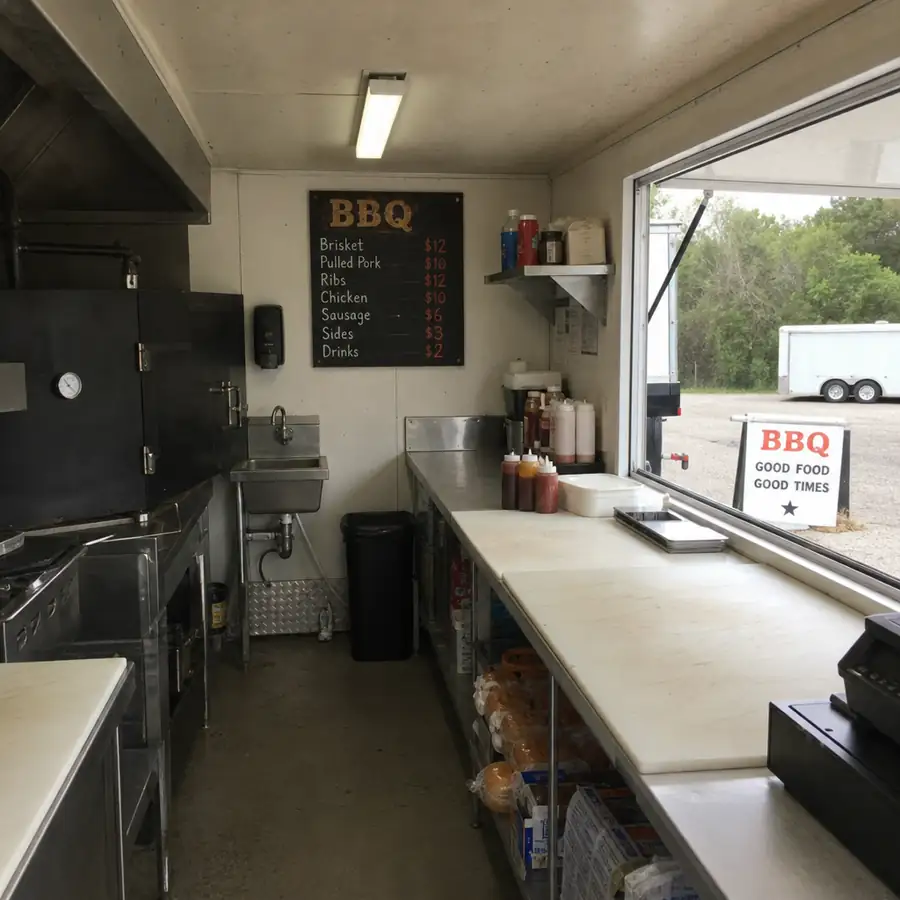 Interior of a BBQ trailer with black smoker, stainless steel prep counters, chalkboard menu, and service window