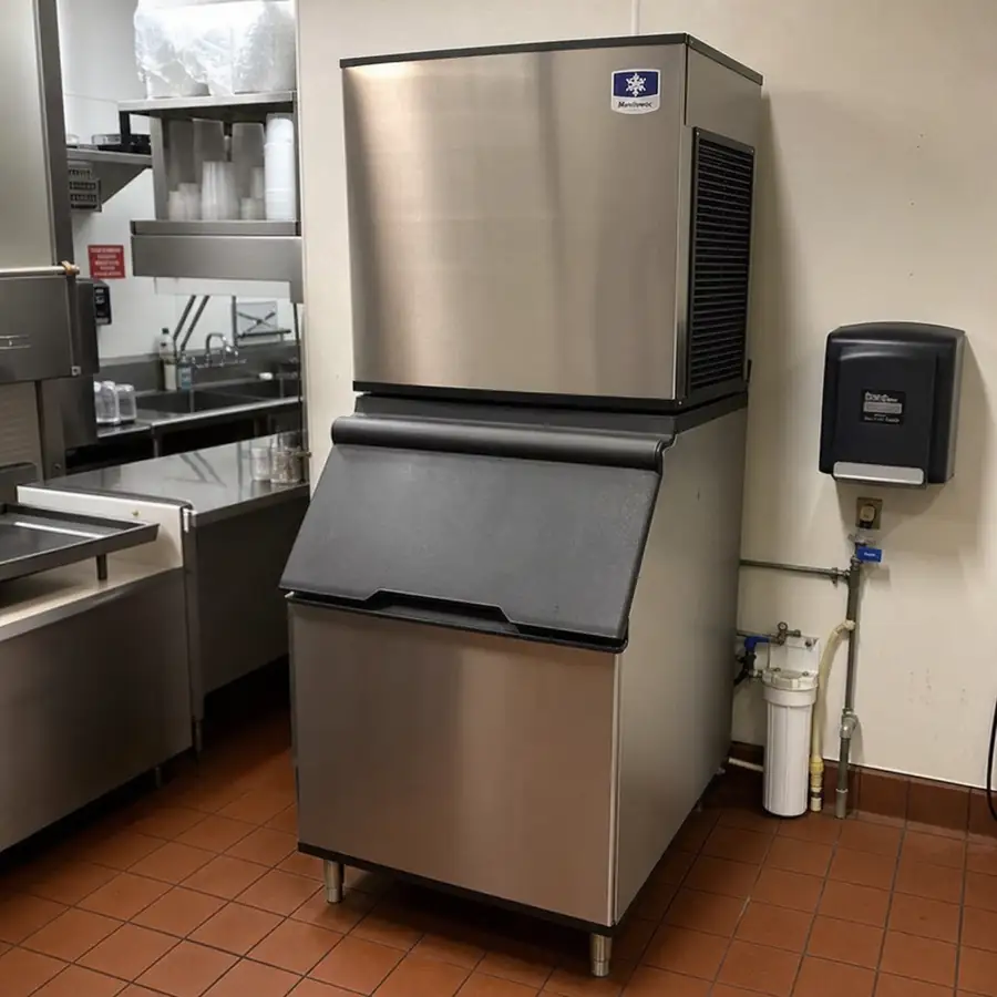 Modular ice machine stacked on a storage bin in a commercial kitchen, with a water filter and supply lines beside it