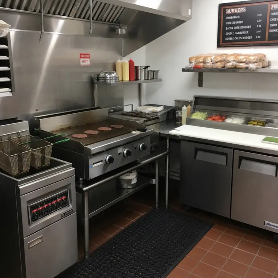 Commercial burger restaurant kitchen with flat-top griddle cooking patties, fryer baskets, refrigerated prep table, and bun storage