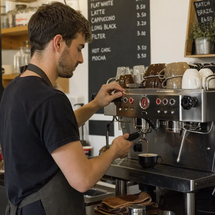 Barista pulling an espresso shot on a commercial machine, with cups on the warmer and a chalkboard menu behind him