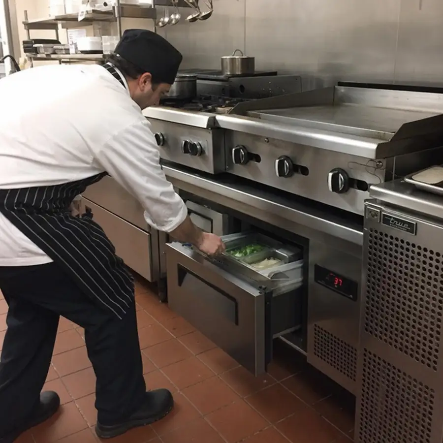 Chef in a white coat and striped apron opening a chef base drawer of ingredient pans beneath a griddle and range