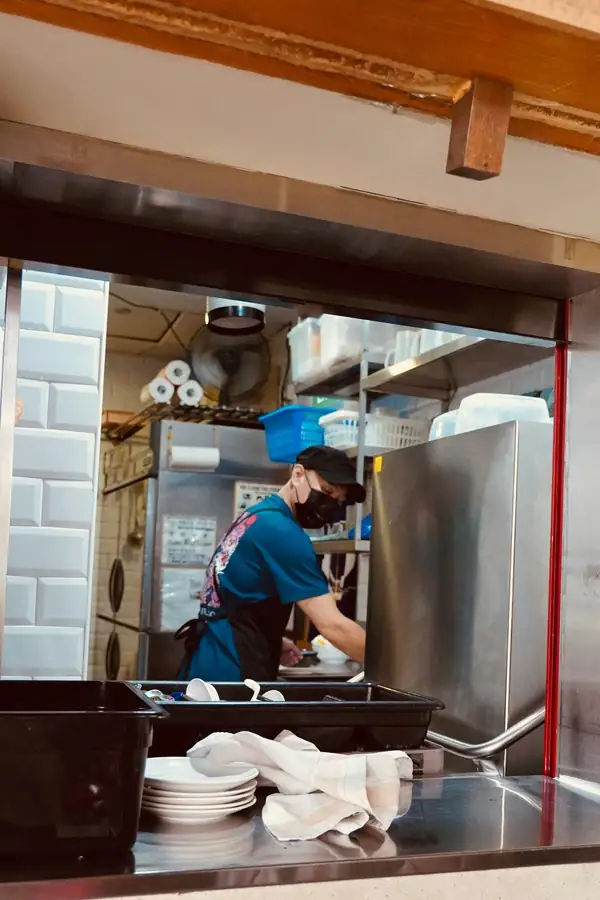 Kitchen worker in a mask and apron preparing food inside a compact stainless-steel food truck kitchen, seen through the service window with stacked plates