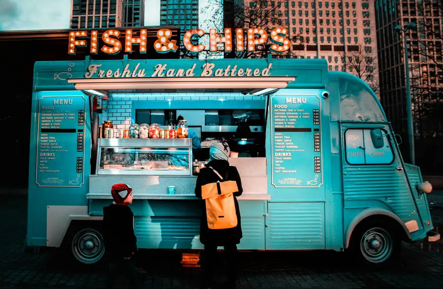 Vintage teal fish and chips food truck with a glowing marquee sign serving customers in a city square at dusk