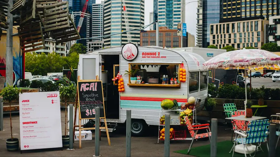 Vintage silver juice trailer named Ronnie Z and the Fabulous Juice in an urban plaza with fruit displays, colorful seating, and city skyscrapers