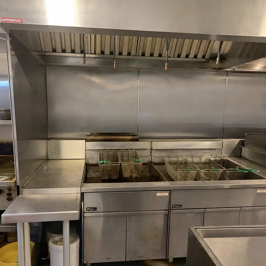 Stainless Pitco commercial fryers under a CaptiveAire exhaust hood, with fry baskets in the oil and a side work table