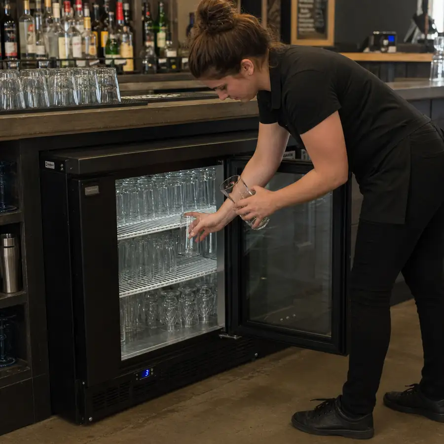 Bartender stocking a black undercounter commercial glass chiller with clean glassware under a bar counter