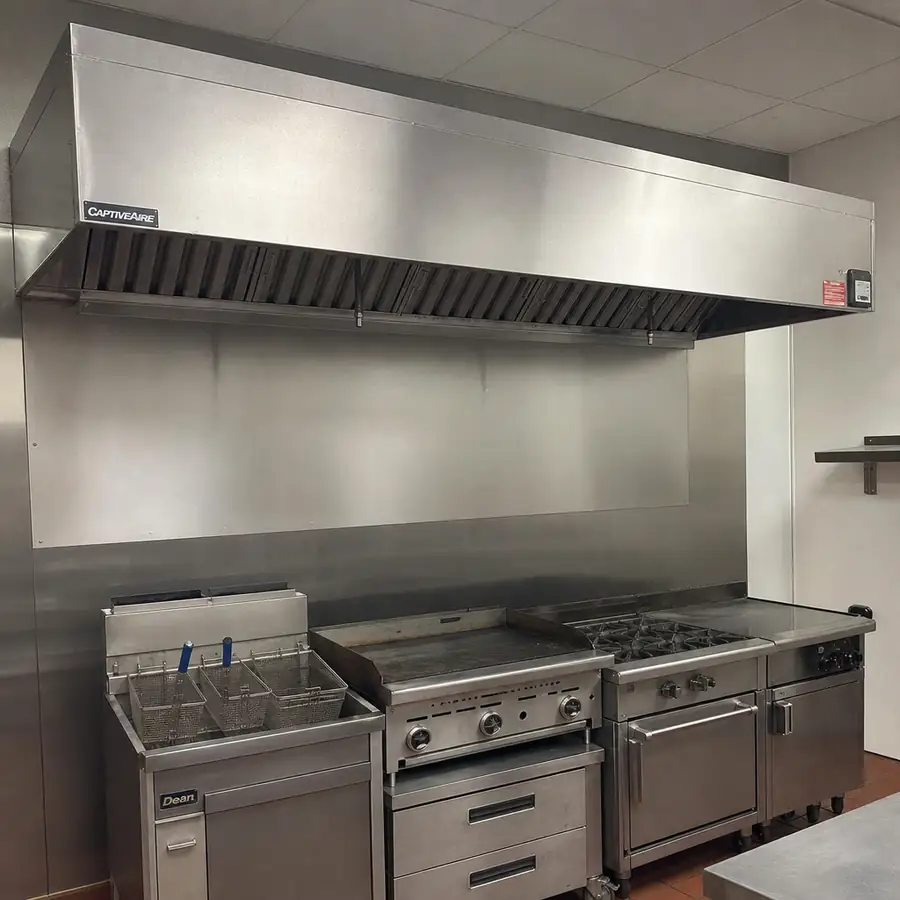 Stainless commercial cook line with an exhaust hood above a fryer, griddle, and gas range with oven