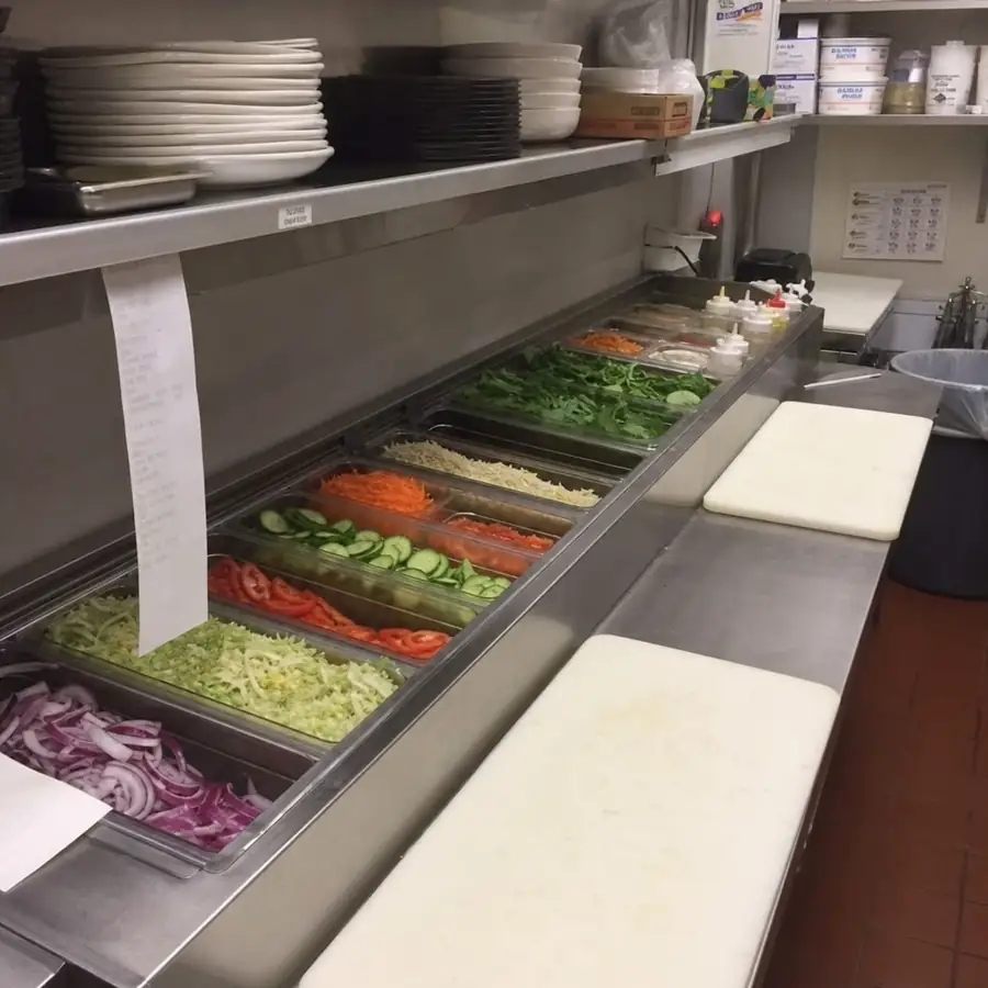 Restaurant kitchen prep station with fresh sliced vegetables in food pans, cutting boards, plates, and condiment bottles on a stainless steel counter