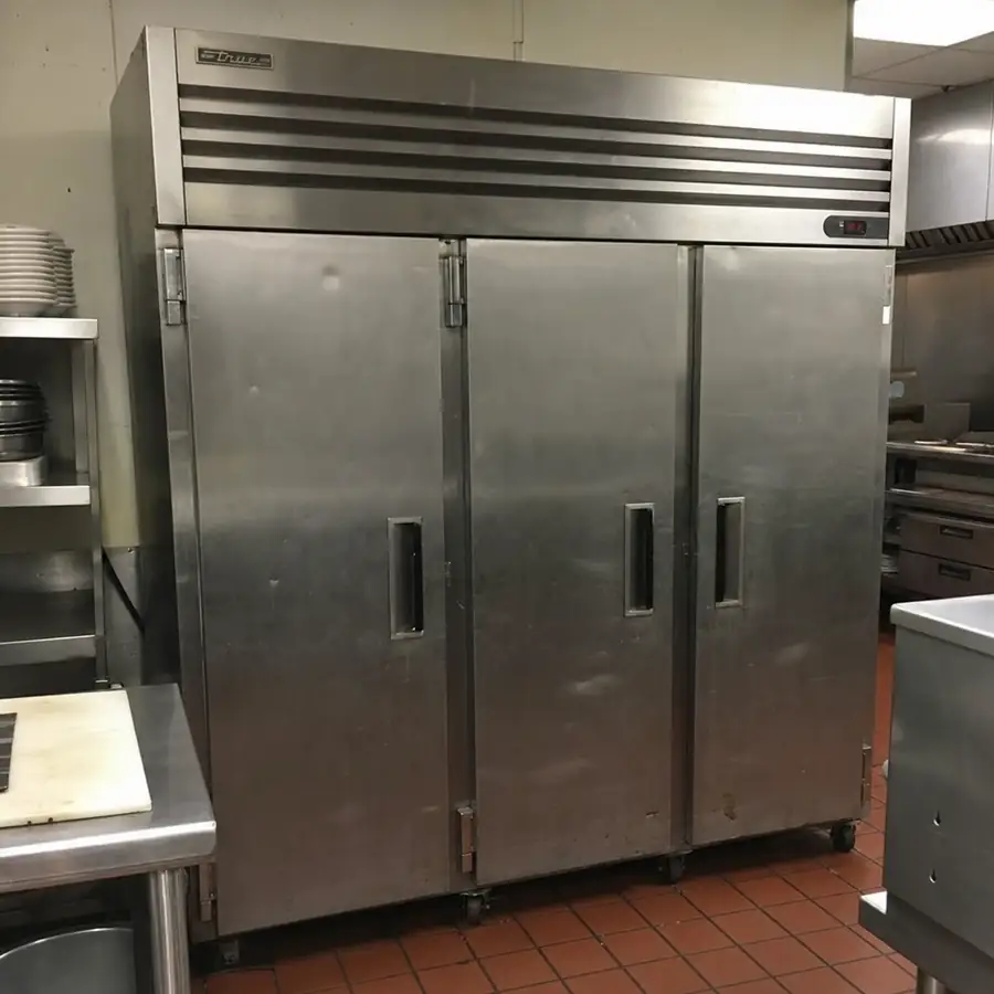Worn three-door commercial reach-in refrigerator with solid doors on casters in a kitchen