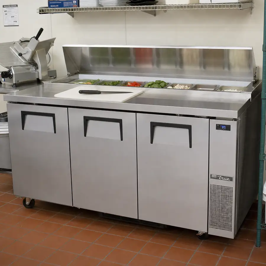 Stainless worktop refrigerator with three doors, a raised rail of ingredient pans, and a cutting board and knife on the prep top